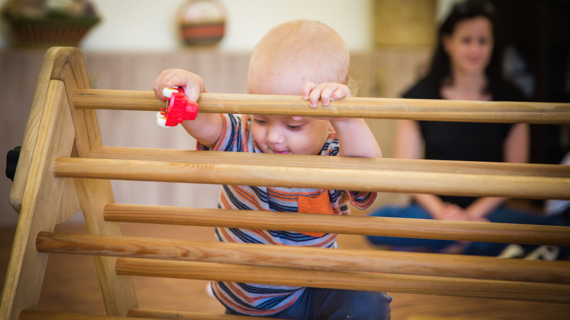 Toddler reaching on a Pikler® triangle with an object in his hand and peering through the slots in the triangle, looking like they may drop the object over the top. 