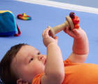Infant laying on back intently inspecting a wooden rattle held in their hands. 