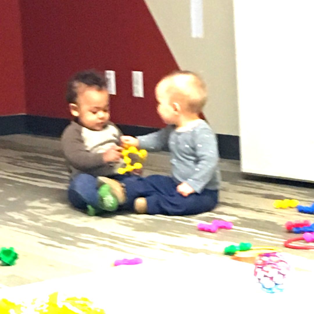 Two babies sitting on a play space floor both holding onto an object while looking at each other.