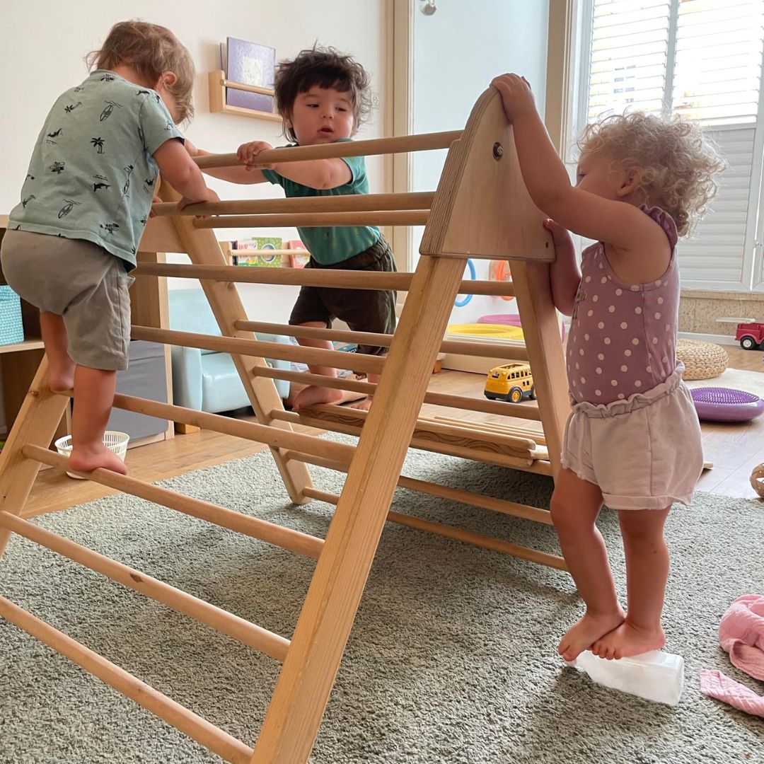 Three toddlers climb on or near a Pikler® Triangle