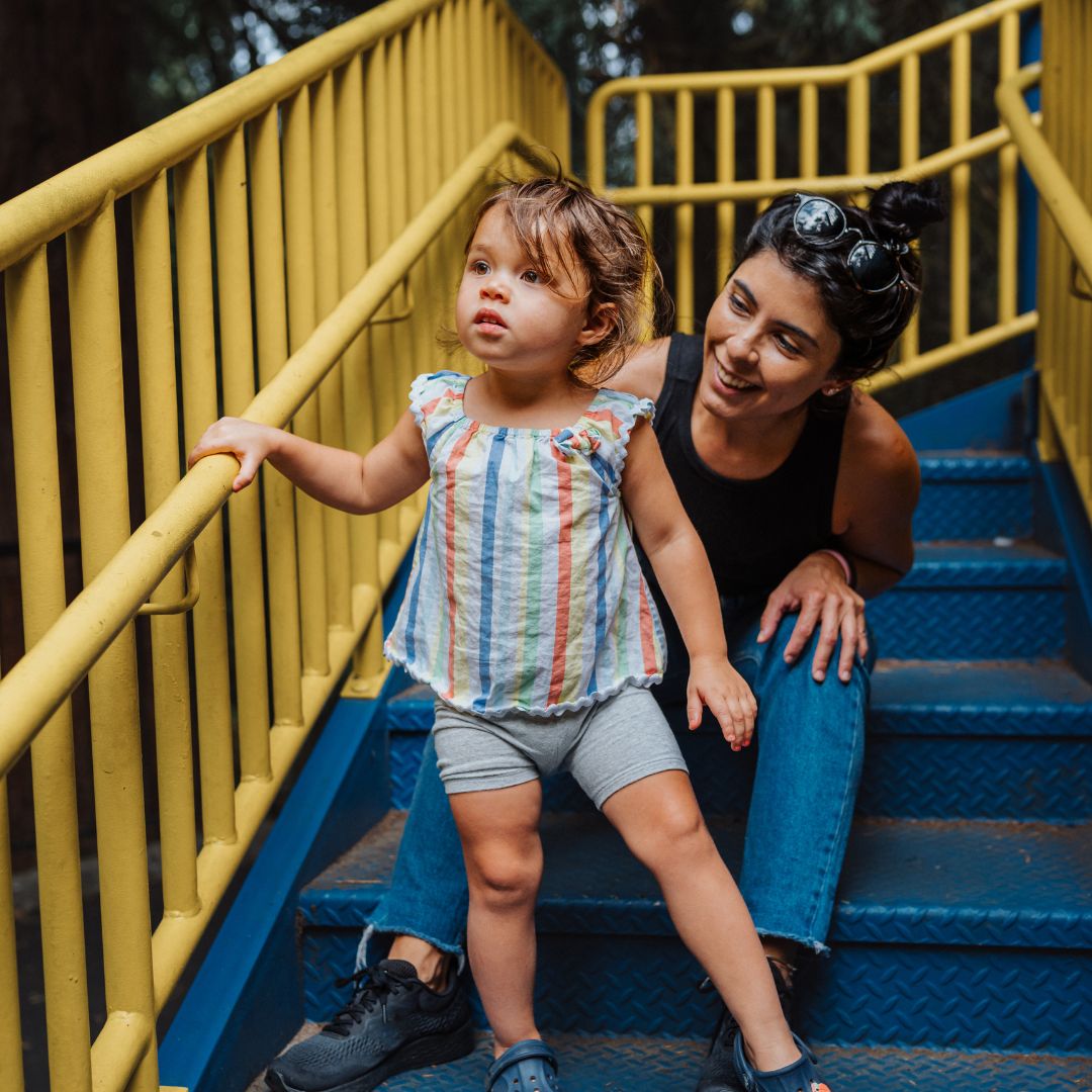 Woman sitting on the stairs to a blue and yellow playground structure smiling at toddler girl who is standing in front of her on the stairs looking off in the distance.