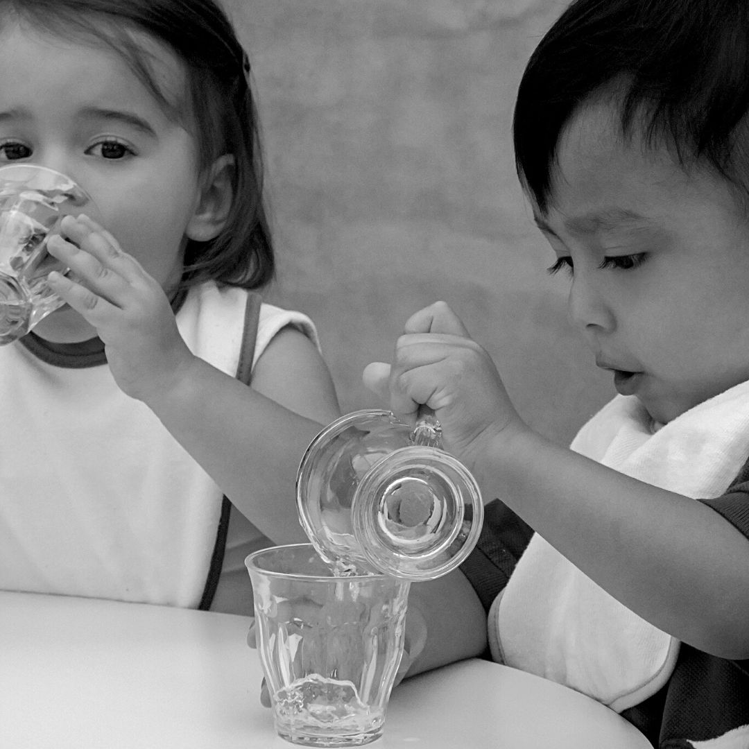 Snack time with toddlers during RIE® Parent Infant Guidance™ Center. One toddler is gently pouring a glass of water from a small glass pitcher into a glass cup.