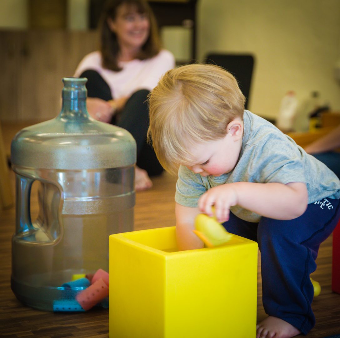 Toddler in foreground putting something into a large yellow cube with large water cooler bottle next to him and adult smiling in the background at a RIE® Parent-Infant Guidance™ Class.