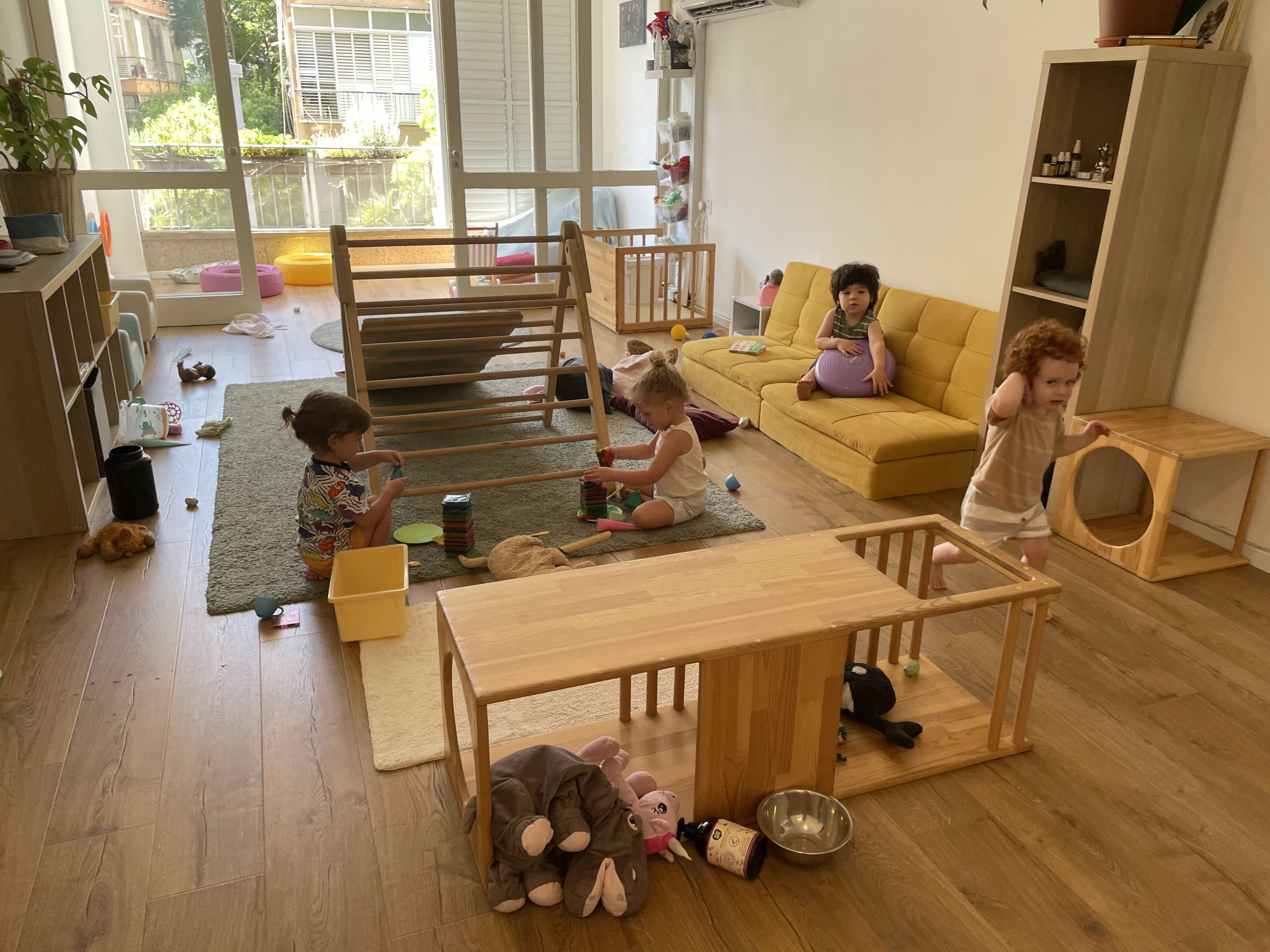 Toddler play space with Pikler® Triangle in the background, climbing cubes in the foreground, and a yellow low cushion sitting area on the side.