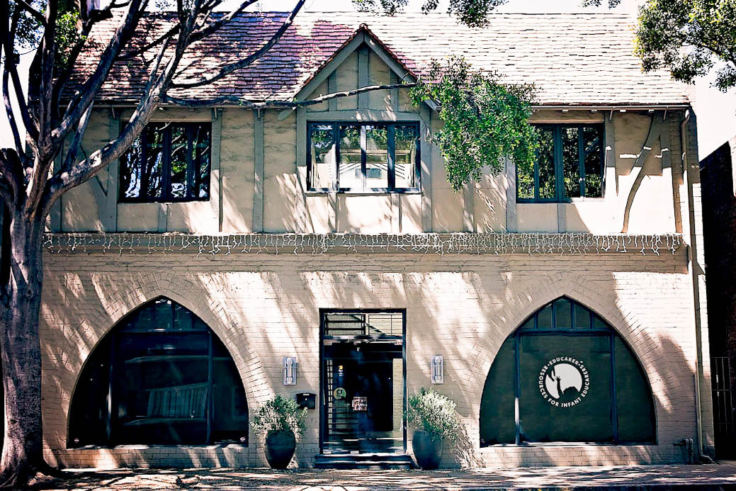 RIE® Pasadena Center with Educaring Logo on right window. It's a light colored two story Tudor building with light dappling on the stucco through the leaves on near by trees.