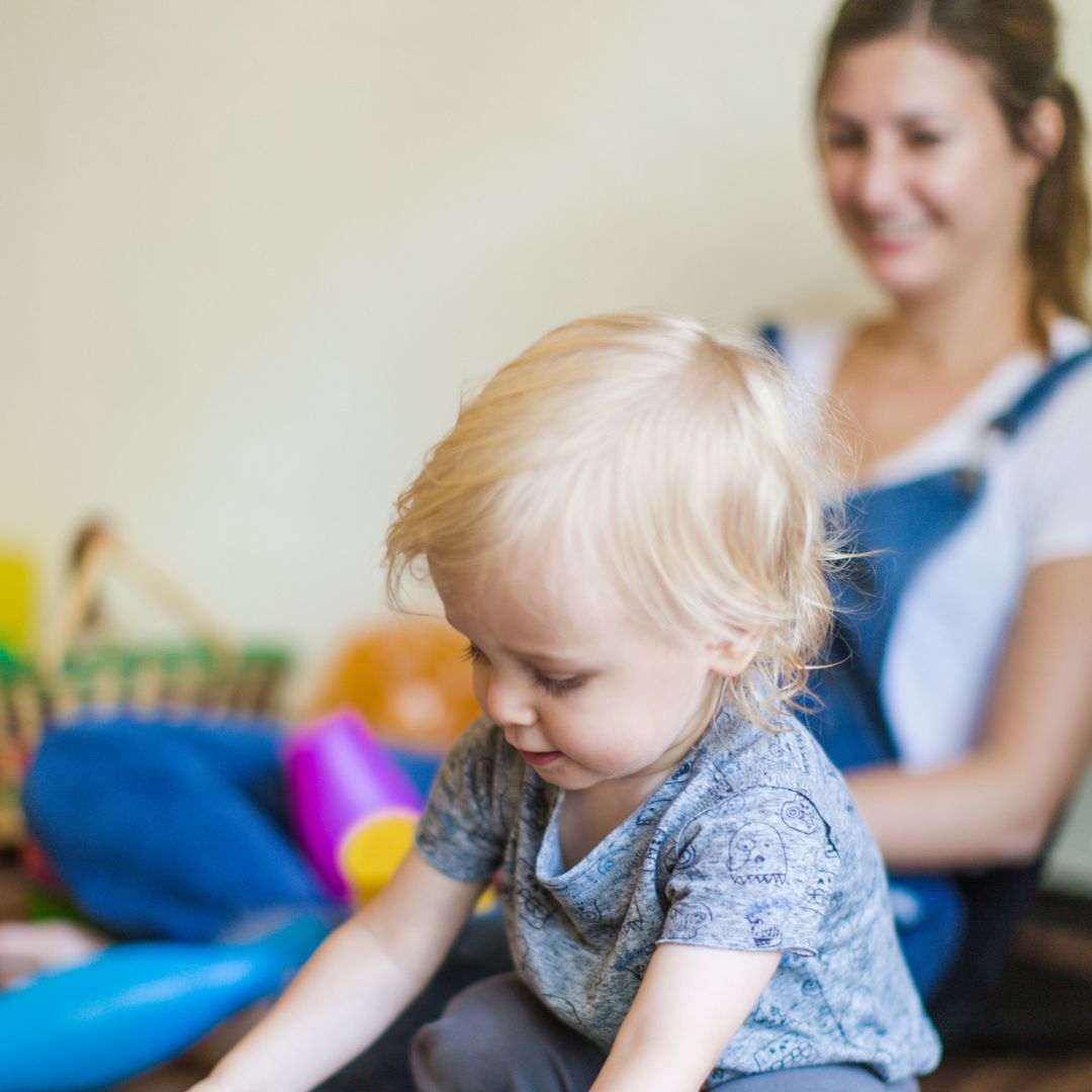 Toddler in foreground with woman in background smiling and looking on at RIE® Parent Infant Guidance™ Class