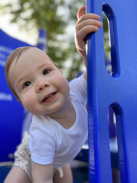 White toddler smiling at the camera while peeking out on top of park play structure.