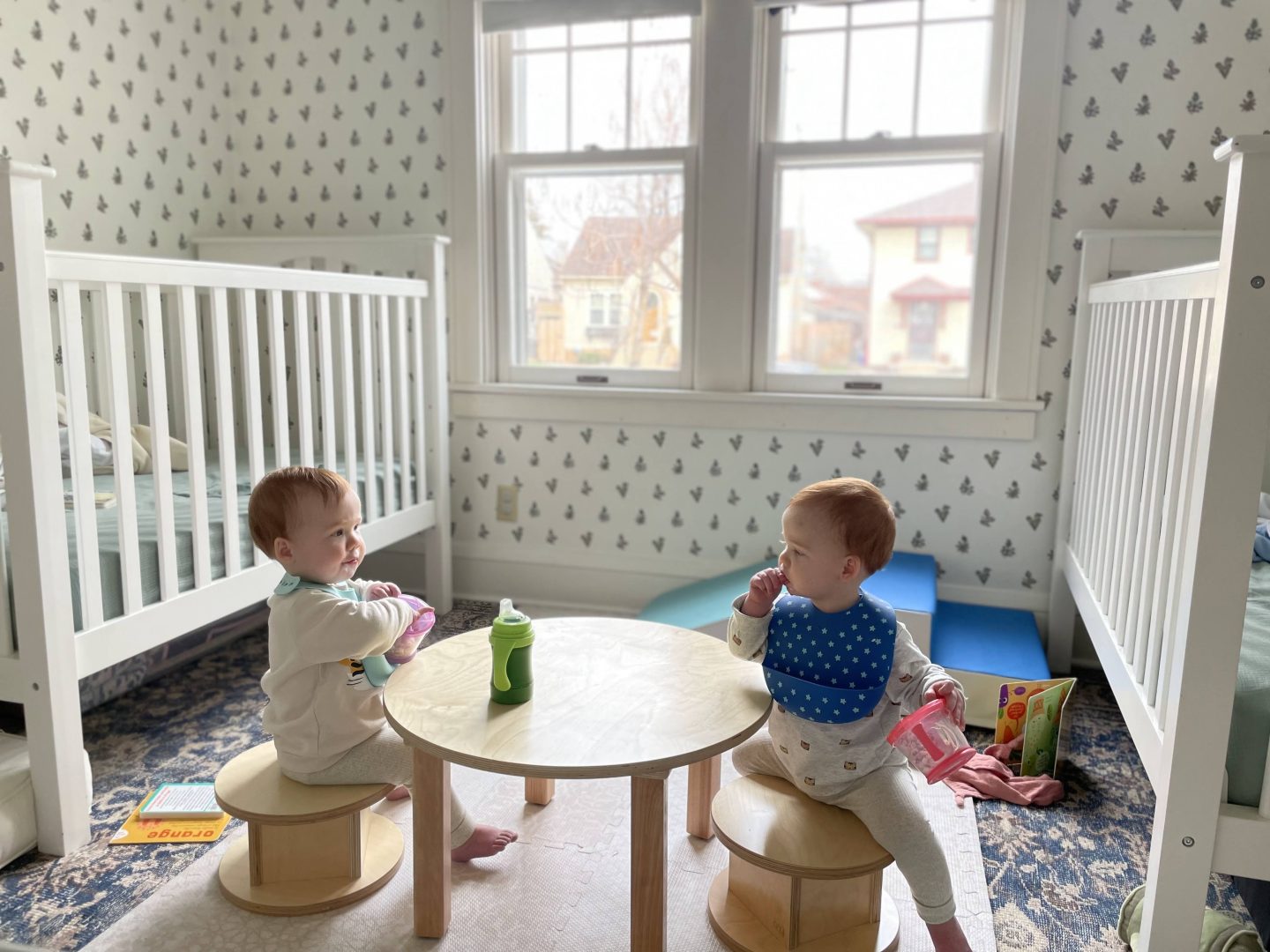 Two toddlers sitting on Magda stools at a low table in their bedroom with two white cribs on either side of the room. They are wearing bibs and having a snack looking at each other.